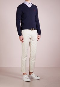 Marineblaue V-Ausschnitt-Strickjacke über einem hellblauen Hemd mit Kragen, kombiniert mit hochgekrempelten weißen Chinos und weißen Sneakers. Hat ein kleines Logo auf der Strickjacke.