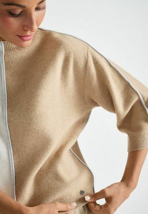 Beige gebreide trui met een hoge kraag, korte mouwen en contrasterende grijze piping. Beschikt over een geribde zoom en een decoratieve knoopdetail.