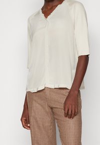 Haut beige clair à manches courtes avec un décolleté en V et des bords festonnés ; tissu doux avec une coupe décontractée, associé à un pantalon à carreaux marron.
