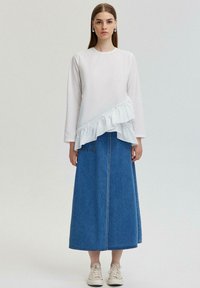 Biely top s dlhými rukávmi a nariaseným detailom, v kombinácii s modrou džínovou maxi sukňou. Model má na sebe béžové tenisky. Jednoduchý, čistý dizajn.