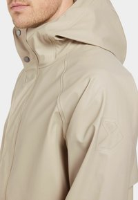 Beige vattentät jacka med huva, tryckknappar och en texturerad logotypbricka på ärmen. Slät, glänsande material med sömnadsdetaljer.