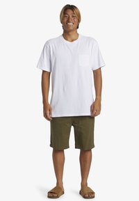 Witte katoenen T-shirt met een borstzak, gecombineerd met olijfgroene shorts en bruine slippers. Model staat in een neutrale pose.