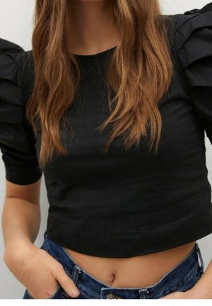 Vrouw met een zwarte cropped top met pofmouwen, lang los haar en blauwe spijkerbroek, handen in de zakken.