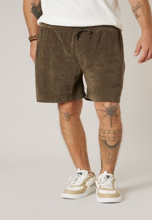 Homme portant un short à cordon de serrage vert olive, des baskets blanches et beiges, avec des tatouages sur les deux jambes et des bracelets au poignet gauche.