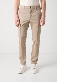 Beige cargobyxor med två sidofickor, tillverkade av ett slätt tyg. Har bälteshällor och knäppning med knapp. Kombineras med vita sneakers.