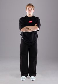 Schwarzes oversize T-Shirt mit rotem Logo, kombiniert mit schwarzen weiten Hosen und weißen Sneakers. Minimalistisches Design und lässige Passform.