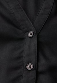 Camicia in tessuto nero con scollatura a V, patta con quattro bottoni scuri e dettagli di cucitura discreti sul davanti.