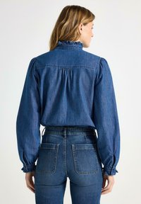 Camicia di denim con colletto alto arricciato, maniche a sbuffo e dettagli arricciati sul retro, abbinata a jeans di denim a vita alta. Colore blu su tutta la superficie.