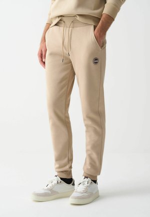 Mann trägt beigefarbene Jogginghose mit Kordelzug und weiße Sneakers, steht mit den Händen in den Taschen vor einem weißen Hintergrund.