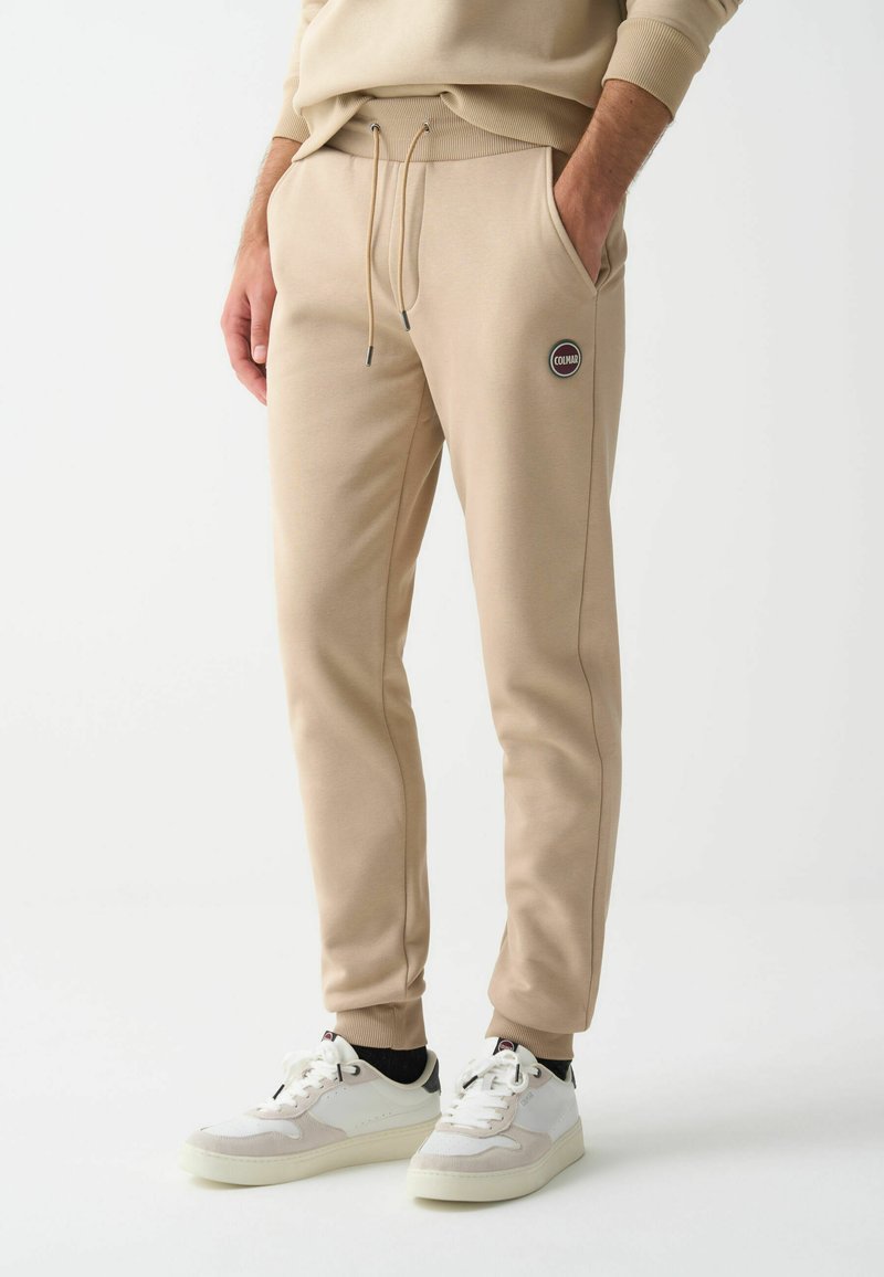 Hombre vistiendo pantalones jogger beige con cintura ajustable y zapatillas blancas, de pie con las manos en los bolsillos sobre un fondo blanco.