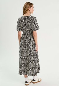 Robe maxi florale noir et blanc avec de courtes manches bouffantes, taille cintrée et silhouette fluide. Le modèle l'a associée à des chaussures sportives et des chaussettes.