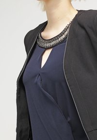 Schwarze Jacke aus strukturiertem Stoff, getragen über einem blauen Oberteil mit einem Ausschnitt in Schlüsselform. Das Oberteil hat einen dekorativen, mit Perlen verzierten Halsausschnitt.