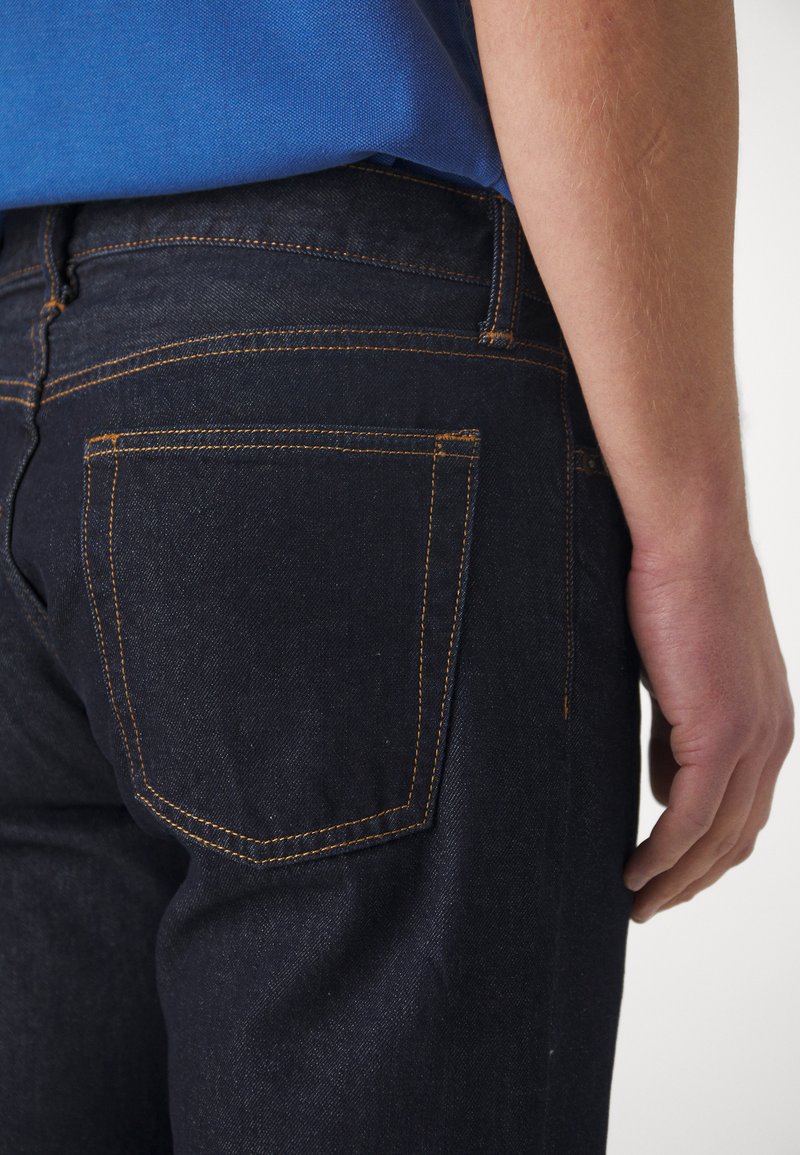 Jeans de mezclilla oscura con costuras naranjas, que presentan un bolsillo trasero con un detalle sutil. La tela tiene una textura suave y un corte clásico.