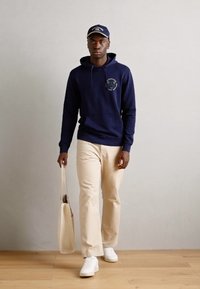 Haut à capuche bleu marine avec un logo, pantalon beige, baskets blanches et un sac fourre-tout léger. Couleurs unies, design décontracté et coupe ample.