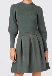 Prążkowany top z długim rękawem w ciemnozielonym kolorze, zestawiony z plisowaną ciemnozieloną spódnicą. Oba elementy mają gładką fakturę i dopasowaną sylwetkę.