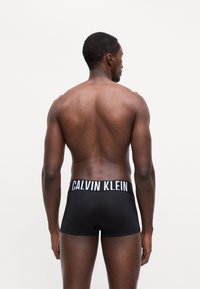 Črne bokserice z belim logotipom Calvin Klein na elastičnem pasu. Gladek material, prilegajoča se oblika, pogled zadaj.