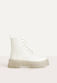 Vit ankelboots i syntetiskt material, med en tjock, genomskinlig sula, snören och en dragflik baktill för enkel på- och avtagning.