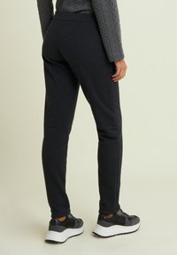 Persona che indossa pantaloni sportivi grigio scuro e un maglione grigio a treccia, abbinati a sneaker bianco e nere, in piedi su uno sfondo neutro.