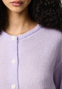 Lila gebreid vest met een ronde hals, geribbelde randen en vier witte knopen aan de voorkant. De textuur is zacht en licht wollig.