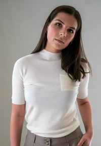 Vrouw met lang bruin haar, gekleed in een witte korte-sleeve trui met een borstzak en een grijze broek, staande tegen een effen lichte achtergrond.