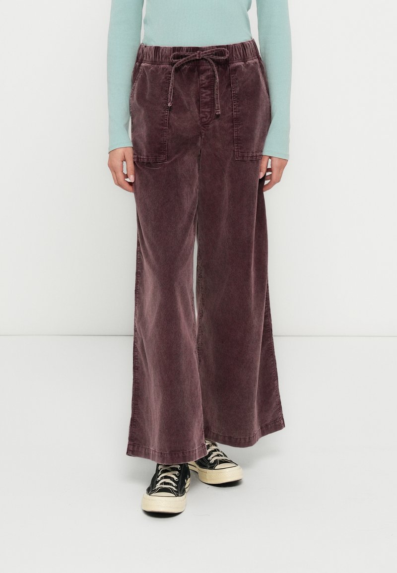 Pantalon large en velours côtelé bordeaux profond avec une ceinture élastique et un cordon de serrage, doté de deux poches avant. Porté avec des baskets noires.