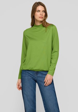 Langärmeliges, hochgeschlossenes Oberteil aus leuchtend grünem Stoff mit lockerem Schnitt und glatter Textur, kombiniert mit hellblauen Jeans mit weitem Bein.