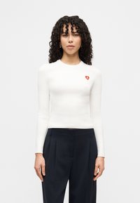 Haut blanc à manches longues côtelé avec un col rond et un petit logo en forme de cœur rouge sur la poitrine, associé à un pantalon sombre et taillé sur mesure.