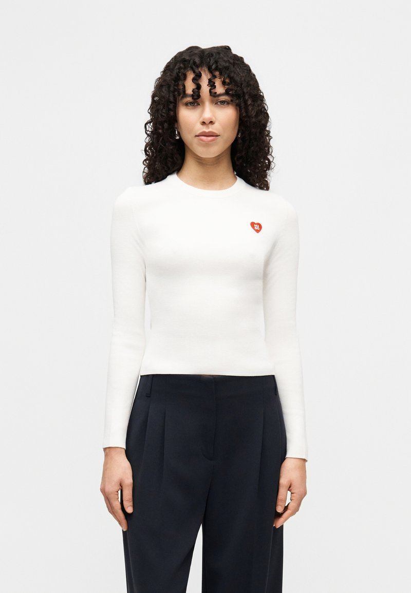 Haut blanc à manches longues côtelé avec un col rond et un petit logo en forme de cœur rouge sur la poitrine, associé à un pantalon sombre et taillé sur mesure.