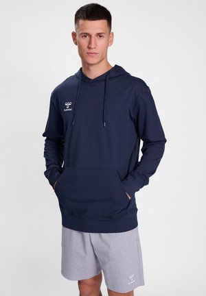 Felpa blu navy con tasca frontale, coulisse regolabile e logo sul petto, abbinata a pantaloni corti grigio chiaro. Texture liscia.