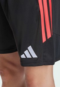 Čierne atletické šortky s tromi červenými vertikálnymi pruhmi, bielym logom Adidas a textúrovanou látkou. Dizajn zahŕňa bočný rozparok pre pohodlie.