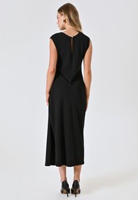 Robe noire midi avec manches courtes, détail de dos à trou de serrure et fente sur le côté. Tissu lisse avec une silhouette fluide. Chaussures à talons hauts noires.
