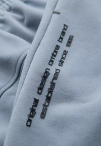 Tissu bleu clair présentant une texture douce avec un texte en relief noir indiquant "design original ombre marque établie depuis 1985."