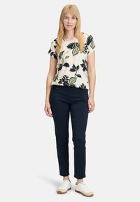 Kremowa bluzka z krótkim rękawem w kwiecisty wzór, z granatowymi i oliwkowymi liśćmi, zestawiona z granatowymi spodniami slim fit i białymi sneakersami z brązowymi podeszwami.