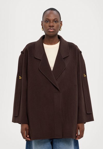 Hnědý vlněný kabát se širokými klopami, dlouhými rukávy a asymetrickým lemem. Obsahuje trojúhelníkový akcent na levém rukávu.