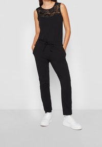 Svart ärmlös jumpsuit med spetsdetaljer vid halslinjen, justerbart dragsnöre i midjan, sidofickor och tapered ben. Bärs med vita sneakers.