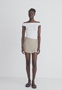 Hvid off-the-shoulder top med rynke detaljer, parret med en beige mini-wrapnederdel med sidelof. Modellen har sorte flade sandaler på.