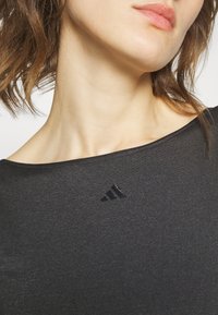 Černý sportovní top s širokým výstřihem, hladký povrch, obsahuje malou černou logo Adidas na hrudi. Detailní záběr horní části těla.