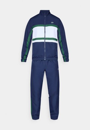Chándal azul marino con chaqueta con cremallera que presenta rayas horizontales blancas y verdes, puños elásticos y pantalones a juego con detalles laterales.