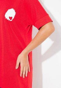T-shirt en coton rouge avec un graphique rond blanc représentant un cœur. Le design a des manches courtes roulées et une coupe décontractée.