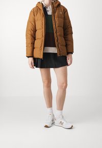 Brown puffer jacket with a hood, textured knit sweater in green, brown, and cream, black pleated skirt, and white sneakers with gold accents.