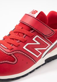 Sneaker New Balance rouge avec sangle velcro, lacets, logo blanc et semelle noire, présenté en gros plan sur un fond blanc.