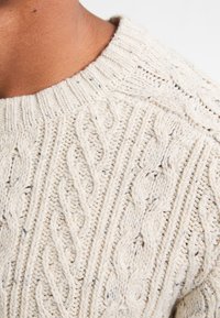 Krämfärgad stickad tröja med kabelstickad mönster, rund halsringning och texturerad yta med subtila prickar.