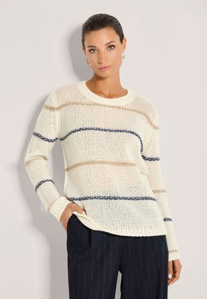 Vrouw met donker haar die een crèmekleurige gebreide trui draagt met horizontale blauwe en beige strepen en donker gestreepte broek, staand tegen een lichte achtergrond.