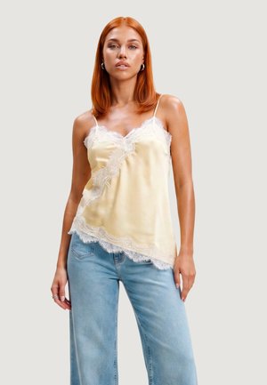 Woman with straight red hair wears a cream lace-trimmed camisole and light blue jeans, standing against a plain light gray background.