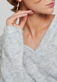 Hellgrauer V-Ausschnitt-Pullover mit weicher, flauschiger Textur und gerippten Bündchen, ergänzt durch einen goldenen Creolen-Ohrring. Hautfarbe am Hals sichtbar.