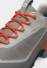 Γκρι και λευκό παπούτσι πεζοπορίας με φωτεινά πορτοκαλί κορδόνια, άνω μέρος από διαπνέον ύφασμα και λογότυπο "SCARPA" στο πλευρικό πέλμα.