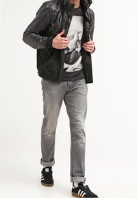 Person som bär en svart jacka, grafisk t-shirt, grå uppvikt jeans och svarta Adidas-sneakers som går mot en enfärgad bakgrund.