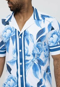 Chemise à manches courtes à boutons avec un motif floral bleu sur fond blanc, avec des accents rayés bleus sur le col et les manches.