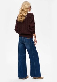 Bordowy sweter robiony na drutach z teksturowanym wzorem, zestawiony z jasnoniebieskimi spodniami w stylu szerokich nogawek. Noszony z beżowymi botkami na obcasie.