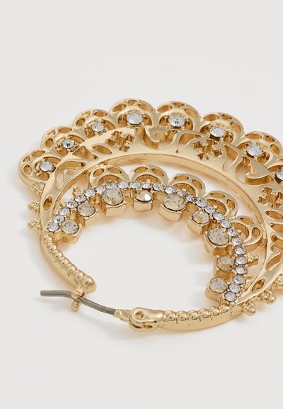 Boucle d’oreille créole dorée avec motifs ajourés complexes et strass clairs disposés en un arc décoratif.